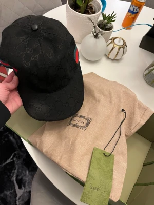 Svart Gucci keps med monogram - Säljer en svart keps från Gucci med diskret GG-monogram över hela tyget och klassiska gröna och röda ränder på sidan. Kepsen har böjd skärm och justerbar rem bak. Kommer med originalpåse och box. Perfekt för dig som vill ha en lyxig och stilren accessoar, en liten missfärgning på kepsen ( svagare svart område på skärmen ) se bild . Solblekt där . 