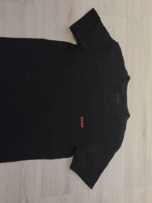Svart t-shirt från Hugo Boss - Klassisk svart t-shirt från Hugo Boss med röd HUGO-logga broderad på bröstet. T-shirten har rund halsringning och korta ärmar. Perfekt basplagg med stilren design som passar till det mesta.