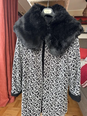 Vändbar svartvit ullkappa - Betsey Johnson - Snygg ullkappa med avtagbar pälskrage. Svartvit leopardmönster på ena sidan och helt svart då man vänder insidan ut. Betsey Johnson. Jackan har rund hals med en stilren metallknäppning framtill. Perfekt för större halsdukar när det blir kallt. Längd från axel och ner ca 82 cm.