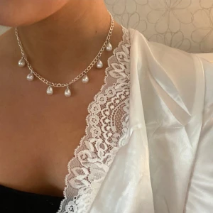 Silverfärgat halsband med pärldroppar - Snyggt halsband i silverfärg med sju hängande pärldroppar. Kedjan är tunn och justerbar, vilket gör att det sitter perfekt som en choker. Pärlorna är vita och ger en elegant touch till din outfit. Perfekt accessoar för att lyfta din stil.