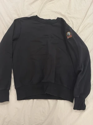Svart sweatshirt från Parajumpers - Svart sweatshirt från Parajumpers med klassisk rund hals och ribbade muddar. På ärmen sitter ett snyggt broderat PJS-märke med texten 'That Others May Live' och 'Anchorage'. Perfekt för en clean och stilren look. ÄKTA