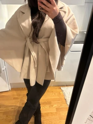 Beige cape-kappa med knytskärp - Snygg beige cape-kappa med bred krage och knytskärp i midjan. Kappan har en lös, flödande passform och är tillverkad i ett mjukt tyg som ger en elegant look. Perfekt för dig som vill ha en trendig och bekväm ytterjacka med modern siluett.