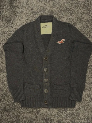 Mörkgrå Hollister Kofta - Säljer denna mörkgrå Hollister koftan. Storlek S och mycket bra skick. Skriv om du har någon fråga. Längd: 60 cm Bredd: 46 cm