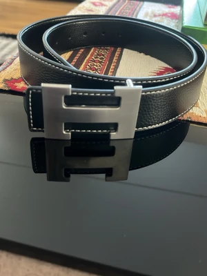 Svart skärp med metallspänne - Snyggt svart bälte med grov struktur och kontrastsömmar. Stort metallspänne i silverfärg med modern design. Perfekt accessoar för att lyfta din outfit.