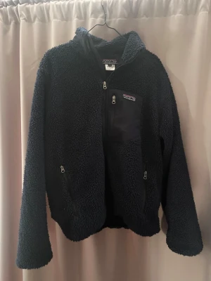 Marinblå teddyjacka från Patagonia - Mysig marinblå teddyjacka från Patagonia med dragkedja framtill, två sidofickor med dragkedja och en bröstficka med logga. Tillverkad i fluffigt material och fodrad med ekologisk bomull. Perfekt för kalla dagar och riktigt skön att ha på sig.