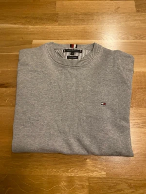 Grå tröja från Tommy Hilfiger - Snygg ljusgrå tröja från Tommy Hilfiger i premium bomull. Klassisk rund halsringning och diskret logga broderad på bröstet. Perfekt basplagg med stilrena detaljer och ribbade muddar vid halsen.