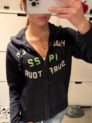 Hollister zip up hoodie huvtröja med dragkedja - Mysig mörkblå zip-hoodie från Hollister med vit och grön text på bröstet.                                     Storlek S.                                                             Modellen har huva med dragsko, två fickor fram och dragkedja. Perfekt för en avslappnad stil och enkel att matcha med jeans.                                              Dragskon är lite sliten annars bra skick!