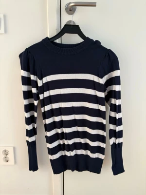 Marinblå och vit randig tröja - Snygg långärmad tröja med breda marinblå och vita horisontella ränder. Tröjan har rund halsringning, puffade axlar och ribbade muddar vid ärmslut och nederkant. Perfekt för en stilren och avslappnad look.