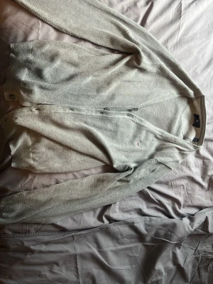 Ljusgrå stickad cardigan - Snygg ljusgrå stickad kofta med knäppning framtill och långa ärmar. Koftan har en diskret liten logga på bröstet och är tillverkad i ett mjukt, tunt material som passar perfekt för lager på lager. Enkel och stilren design som funkar till det mesta.