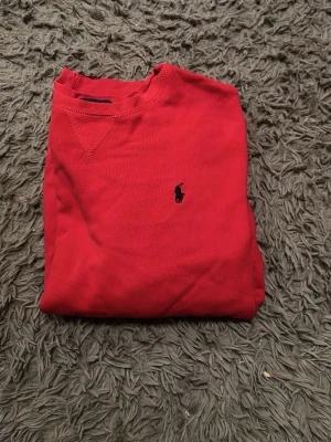 Röd crewneck från Ralph Lauren - Snygg röd crewneck från Ralph Lauren med klassisk blå logga broderad på bröstet. Tröjan har rund halsringning och är tillverkad i mjukt bomullsmaterial. Perfekt för en clean och stilren look.