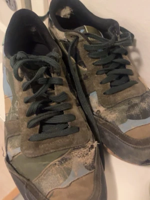 Valentino camo sneakers med nitar - Säljer ett par unika Valentino sneakers med camomönster i grönt, beige och blått. Skorna har svarta skosnören, mockadetaljer och coola nitar på hälen. Perfekta för dig som gillar streetwear och vill sticka ut med exklusiva detaljer.