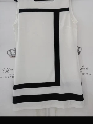 Vit klänning med svarta ränder - Stilren ärmlös vit klänning med svarta grafiska ränder framtill och två svarta ränder nedtill på baksidan. Klänningen har rak passform och rund halsringning. Perfekt för dig som gillar minimalistisk och modern design.