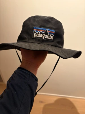 Svart solhatt från Patagonia - Säljer en svart solhatt från Patagonia med bred skärm och justerbart hakband. Hatten har den klassiska Patagonia-loggan framtill med färgglada detaljer. Perfekt för soliga dagar och outdoor-äventyr.