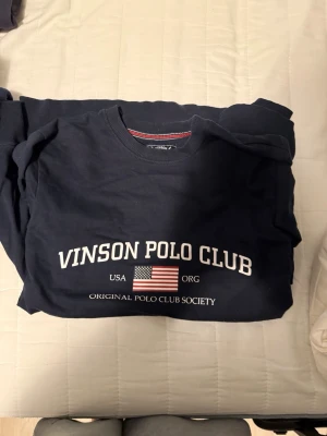 Mörkblå sweatshirt Vinson Polo Club - Mörkblå sweatshirt från Vinson Polo Club med rund hals och tryck framtill i vitt samt amerikansk flagga. Klassisk college-stil med ribbade muddar vid ärmslut och hals. Perfekt för en avslappnad och sportig look.