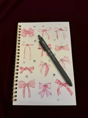 Anteckningsbok med rosettmotiv & penna - Säljer en spiralbunden anteckningsbok med vit pärm dekorerad med rosa rosetter och glittriga stenar. Medföljer en grå penna med texten 'Dream BIG AND Work HARD' i vitt. Perfekt för skolarbete, dagbok eller kreativa idéer.