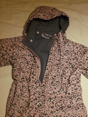 Rosa leopardmönstrad overall  - Jättefin overall med rosa leopardmönster och grått fleecefoder. Overallen har huva, dragkedja och kardborrband framtill. Perfekt för regniga dagar och ger en cool look med sitt djurmönster. Storlek 98/104
