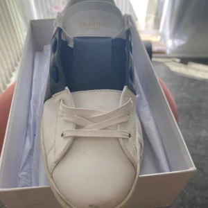 Valentino Open sneakers vit/blå - Snygga sneakers i vitt skinn med bred blå läderdetalj över mitten. Klassisk rund tå, vita snören och ikoniska nitar på sulan. Perfekta för dig som gillar stilrena och lyxiga sneakers med en twist.