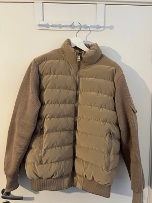 Beige moncler pufferjacka med stickade ärmar - Snygg beige pufferjacka med quiltad front och rygg samt stickade ärmar och muddar. Jackan har hög krage, två dragkedjefickor fram och en ärmficka. Perfekt för kalla vinterdagar och ger en modern look. Nypris 4-8tusen så skriv så diskuterar vi priset.