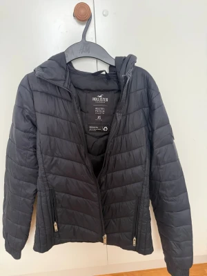 Svart pufferjacka från Hollister XS - Snygg svart pufferjacka från Hollister med huva och dragkedja framtill. Jackan har två fickor med dragkedja och är tillverkad av återvunnet material. Perfekt för kalla dagar eller nu till våren  och ger en clean, stilren look. 