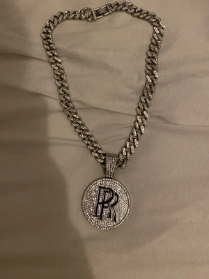 Kedjehalsband med R-berlock - Snyggt halsband i silverfärg med grov kedja och en rund berlock täckt av glittrande stenar. Berlocken har ett stort bokstaven R i mitten, vilket ger en personlig touch. Perfekt accessoar för dig som vill sticka ut och gillar bling.