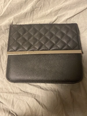 Svart quiltad kuvertväska med metallist - Svart handväska. Magneten är halvt av men är fortfarande på så den går att stänga. Finns även ett snöre man får med som hör till väskan så man kan ha den runt halsen (kan skicka mer bilder om man är intresserad)