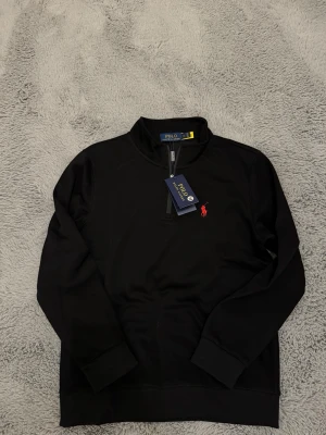 Svart quarter zip från Polo Ralph Lauren - Snygg svart quarter zip från Polo Ralph Lauren med klassisk röd logga broderad på bröstet. Tröjan har hög krage, dragkedja framtill och ribbade muddar vid ärmar och nederkant. Perfekt för en clean och stilren look. ⚠️⚠️Den är dock liten i storlek så skulle rekommendera tröjan till någon som bär XS/S. (160-170)⚠️⚠️