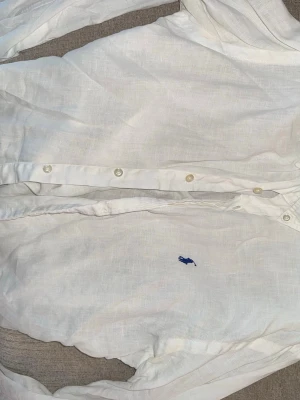 Vit linneskjorta från Ralph Lauren - Klassisk vit skjorta från Ralph Lauren i linne med blå broderad logga på bröstet. Skjortan har knappar framtill och smal passform. Perfekt för en clean och stilren look.