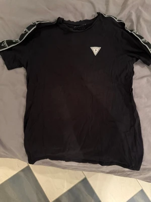 Svart t-shirt från Guess med logga - Svart t-shirt från Guess med vita loggor längs axlarna och en liten trekantig Guess-logga på bröstet. Klassisk rund hals och korta ärmar. Tillverkad i mjukt material som känns skönt mot huden. Perfekt för en stilren och sportig look.
