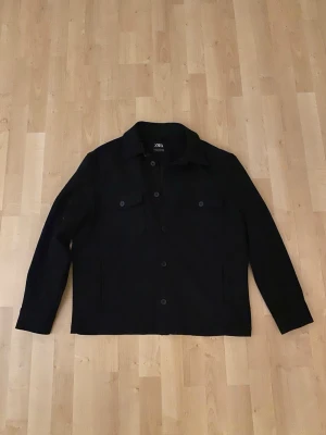 Svart overshirt från Zara - Stilren svart overshirt från Zara med klassisk krage och knäppning framtill. Två bröstfickor med lock och knappar samt raka ärmar med knapp vid ärmslut. Perfekt lager-på-lager-plagg i ett mjukt material. Säljer då jag köpte en för liten storlek. Jag är 190 cm lång och bredaxlad.