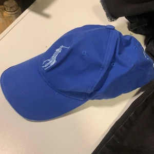 Blå keps från Ralph Lauren - Snygg blå keps från Ralph Lauren med klassisk broderad polospelare framtill och siffran 3 på sidan. Justerbart spänne bak och broderad text. Perfekt accessoar för en sportig och avslappnad stil.