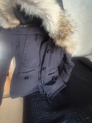 Svart parkas från Canada Goose - Snygg svart parkas från Canada Goose,äldre modell.med stor luva och fluffig pälsdetalj. Jackan har flera fickor med knappar och dragkedjor, samt den klassiska röda loggan på ärmen.