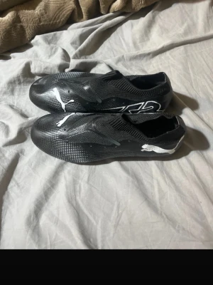 Svarta Puma fotbollsskor - Säljer ett par svarta fotbollsskor från Puma med vit logga på sidan. Skorna har en strömlinjeformad design och ovandel i syntetmaterial som ger bra bollkontroll. Lätta och smidiga för snabba rörelser på planen. Perfekta för dig som vill ha stilrena och funktionella skor.