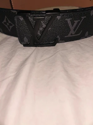 Svart Louis Vuitton bälte med monogram - Snyggt svart bälte från Louis Vuitton med klassiskt LV-monogram i grått och stor LV-logga på spännet. Bältet är gjort i skinn och har en exklusiv känsla med detaljer i mörka toner. Perfekt accessoar för att lyfta din outfit.