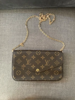 Louis Vuitton monogram kuvertväska - Säljer en brun kuvertväska från Louis Vuitton med klassiskt monogrammönster och guldfärgad kedja. Väskan har en elegant, rektangulär form och stängs med en knapp framtill. Materialet är tåligt canvas med detaljer i guldmetall. Perfekt för dig som gillar lyxig och tidlös design.
