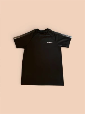 Svart McKenzie t-shirt med tryck - Snygg svart t-shirt från McKenzie med vitt tryck på bröstet och diskreta grå detaljer längs axlarna. Klassisk passform och rund halsringning, perfekt för en sportig look. Tillverkad i mjukt och lätt polyestermaterial.