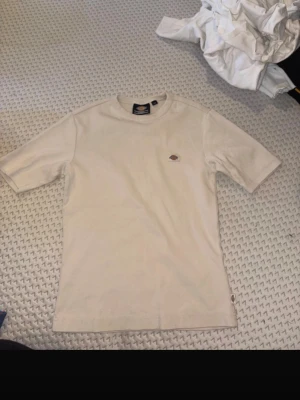 Beige t-shirt från Dickies - Säljer en stilren beige t-shirt från Dickies med klassisk logga på bröstet. T-shirten har rund halsringning och korta ärmar. Tillverkad i mjuk bomull som är skön att bära. Perfekt till jeans eller shorts.