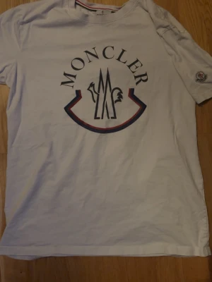 Vit Moncler t-shirt med stor logga - Vit t-shirt från Moncler med stor tryckt logga i svart, rött och blått på bröstet. Klassisk rund halsringning och korta ärmar. Liten Moncler-patch på ena ärmen. Perfekt för dig som gillar exklusiva streetwear-plagg.