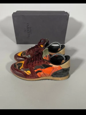 Valentino camo sneakers i rött och orange - Unika sneakers från Valentino med coolt kamouflagemönster i rött, orange, svart och beige. Skorna har låga skaft, vinröda mockadetaljer och snörning. Sulan är beige och ovandelen är en mix av mocka och textil. Perfekta för dig som vill sticka ut.