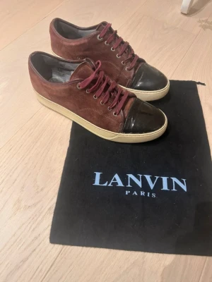 Lanvin vinröda sneakers med mocka - Snygga sneakers från Lanvin i vinröd mocka med mörkbruna tåpartier i läder och matchande vinröda skosnören. Skorna har en beige sula och klassisk låg profil. Perfekta för dig som gillar exklusiva detaljer och stilren design.