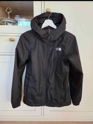 Svart vindjacka The North Face XS - Svart vind/skal/regnjacka från The North Face med huva, dragkedja och två sidofickor. Tillverkad i lätt, vindtätt material med elastiska muddar vid ärmsluten. Logga på bröstet och enkel, stilren design som funkar till det mesta. Använd max 5 ggr. Nyskick. Inökp hösgen 2025, säljes pga fel storlek. Svart i färgen.  Nypris: 1449 kr.