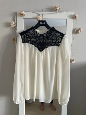 Vit blus med svart spetsdetalj - Snygg vit blus med lång ärm och svart spets upptill som ger en elegant kontrast. Blusen har rund halsringning och knäppning bak i nacken. Materialet är tunt och flowy, perfekt för en chic look. Passar till både jeans och kjol.
