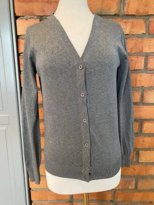 Stadium Grå kofta strl 40 - En enkel och stilren grå kofta med v-ringning och knäppning framtill. Koftan har långa ärmar, ribbade muddar och är tillverkad i ett mjukt, tunt stickat material. Perfekt att slänga över en t-shirt eller linne.