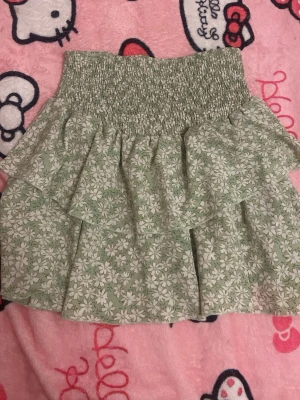 Grön blommig volangkjol från SHEIN - Söt grön kortkjol från SHEIN med vitt blommönster och dubbla volanger. Kjolen har en bred, smockad midja som ger en bekväm och flexibel passform. Perfekt för vår och sommar när du vill ha en lätt och luftig stil.