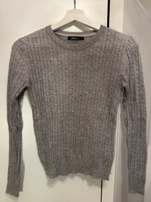 Grå kabelstickad tröja Gina Tricot - Snygg grå kabelstickad tröja från Gina Tricot med rund halsringning och långa ärmar. Tröjan har ett klassiskt mönster och ribbade muddar vid ärmslut och nederkant. Perfekt att matcha med jeans eller kjol för en trendig look.