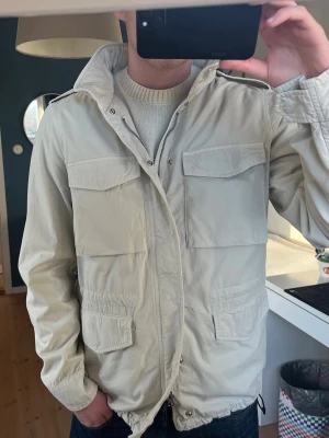 Beige vindjacka från Aspesi - Helt oanvänd 10/10 skick. Stilren beige fieldjacka från Aspesi med fyra stora fickor framtill och hög krage med kardborrestängning. Jackan har dragsko i nederkant och axelklaffar. Tillverkad i lätt material som passar perfekt till vår och höst. Pris kan diskuteras. 