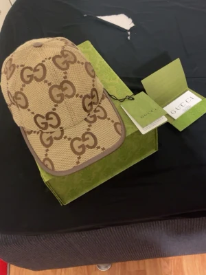 Beige keps från Gucci med GG-mönster - Snygg beige keps från Gucci med det klassiska GG-monogrammet i brunt över hela kepsen. Justerbar rem baktill för perfekt passform. Tillverkad i canvasmaterial med bruna detaljer runt skärmen och kanten. Perfekt för dig som vill ha en exklusiv och trendig accessoar.