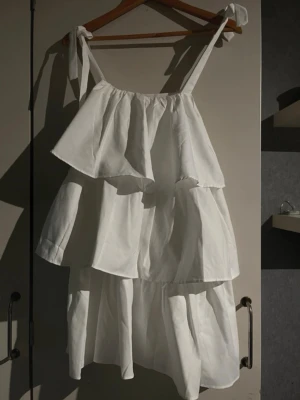 Skolavslutningsklänning - Söt vit klänning med flera volanger och knytbara axelband. Klänningen har en luftig passform och är perfekt för varma dagar. Tillverkad i ett lätt tyg som ger en skön känsla. Stilren och enkel design som känns modern och fräsch.