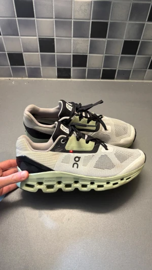 On Running Cloudmonster sneakers - Snygga On Running Cloudmonster sneakers i beige och svart med mesh-ovandel och gröntonad chunky sula. Skorna har svarta snören, ventilerande material och cool design med On-logga på sidan. Perfekta för dig som gillar streetwear och sportig stil.