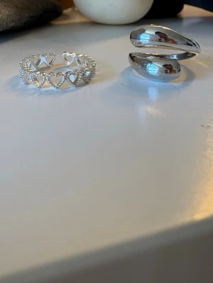Två silvriga ringar med hjärtdetaljer - Säljer två snygga ringar i silverfärg. Den ena ringen har öppna hjärtan runt hela bandet och små glittriga detaljer, medan den andra är bredare och har en modern, spiralformad design. Perfekta accessoarer för att lyfta din stil.