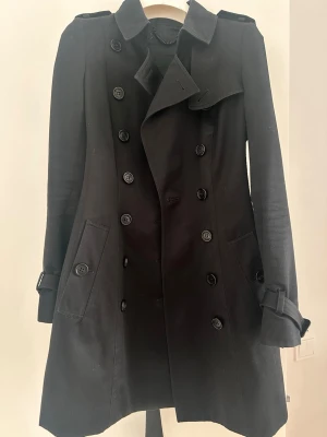 Svart klassisk trenchcoat med bälte - Stilren svart trenchcoat med dubbelknäppning, axelklaffar och bälte i midjan. Jackan har två snedställda fickor och dekorativa spännen vid ärmsluten. Perfekt för dig som vill ha en tidlös och snygg look. Materialet känns kraftigt och håller formen.